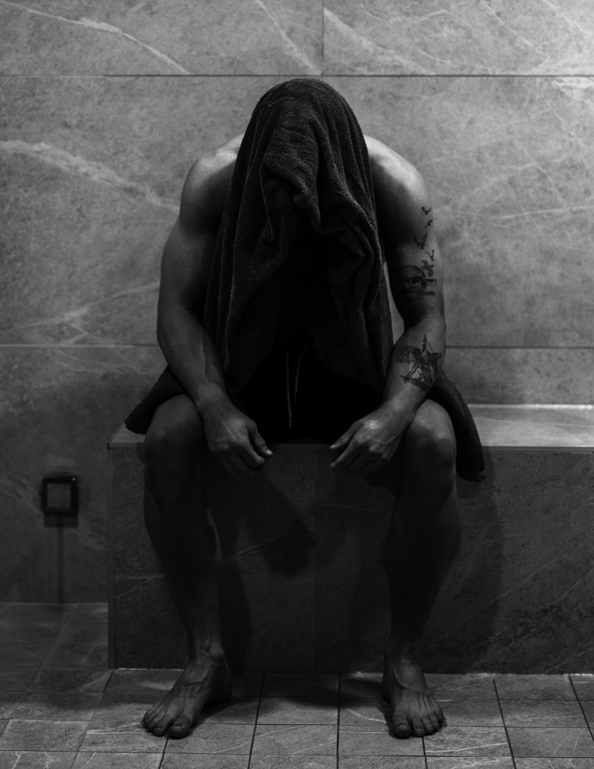 Anthony in black and white, seated on a tiled bench with a towel over his head