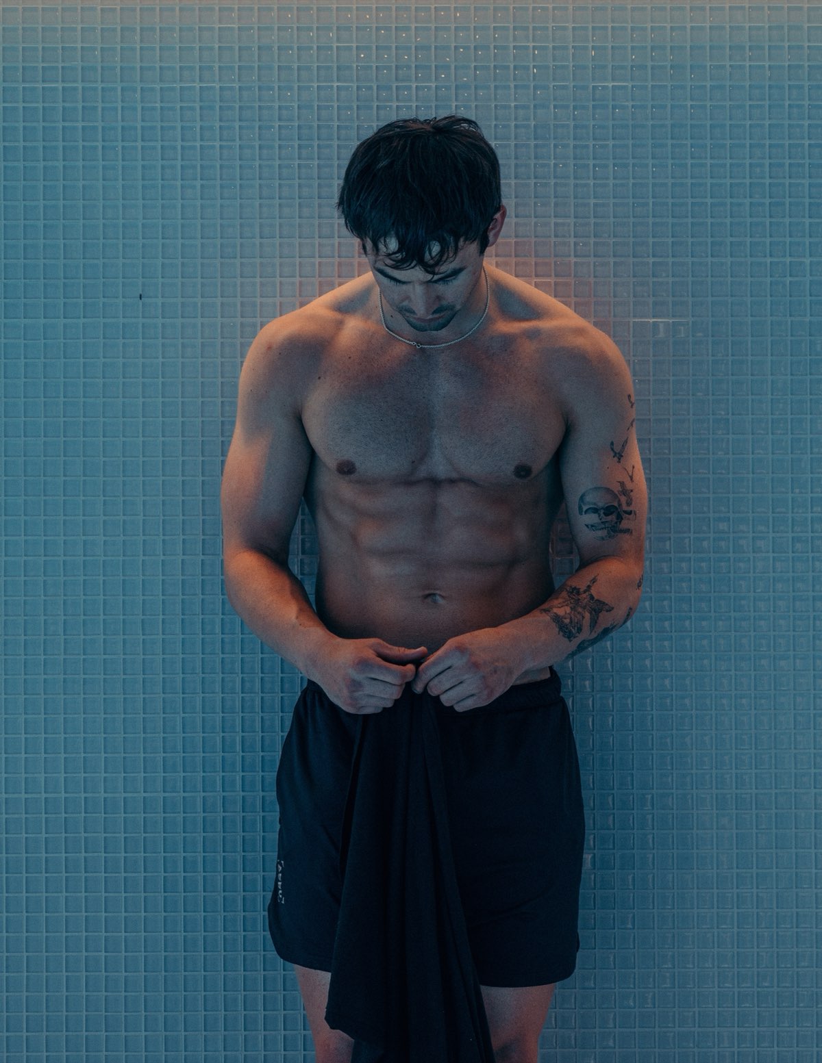 Anthony standing against a blue tiled wall, towel in hand, looking down