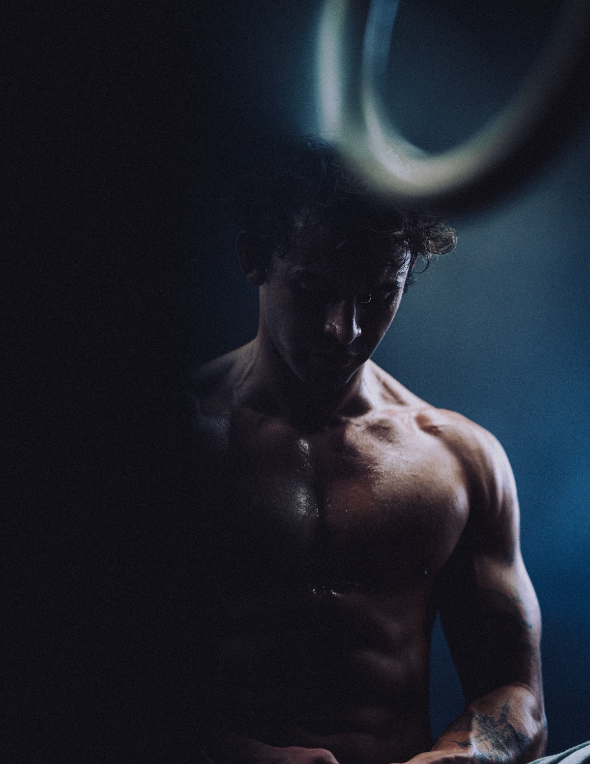 Anthony shirtless in low light, looking down, a blurred gymnastic ring above his head