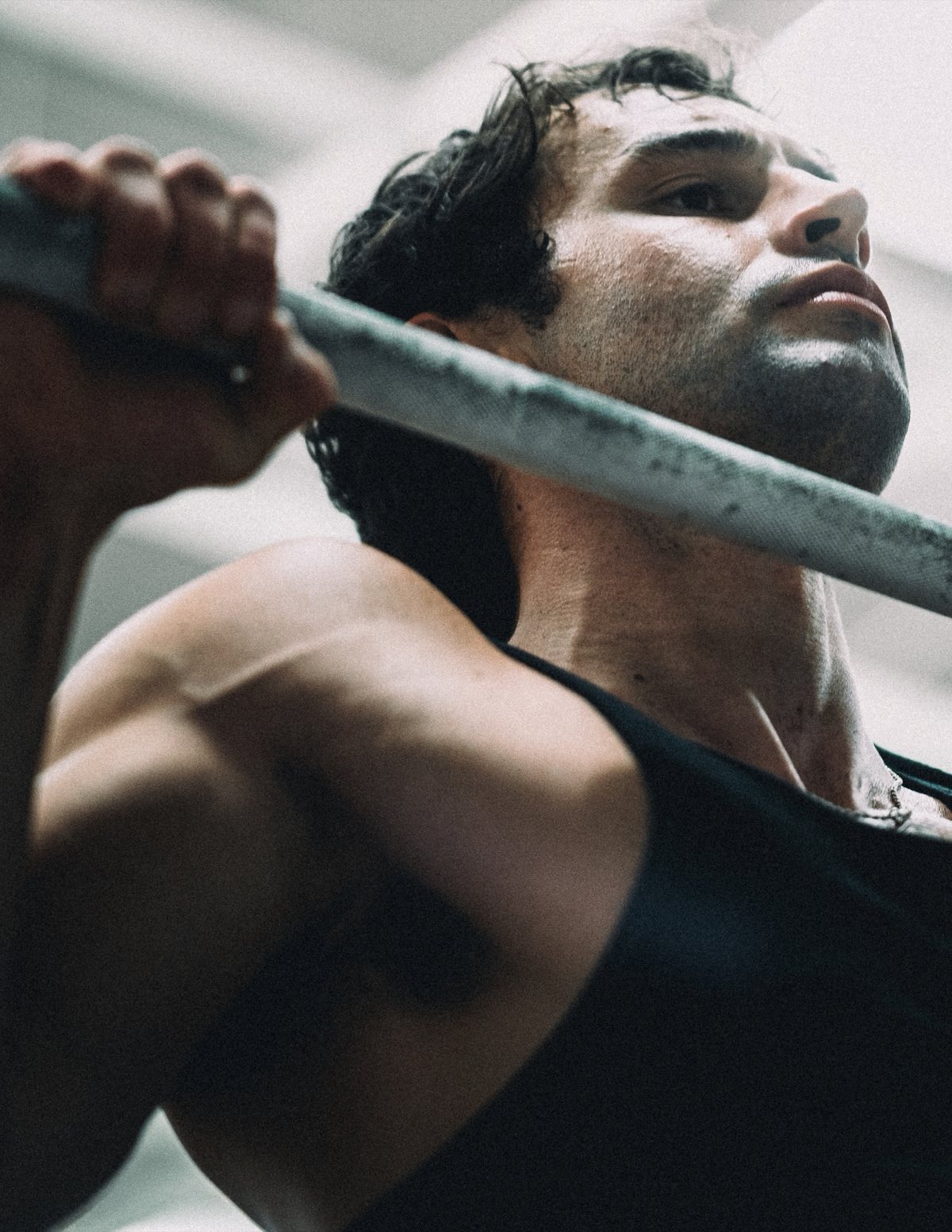 Anthony gripping a bar overhead, looking up, in a black tank top
