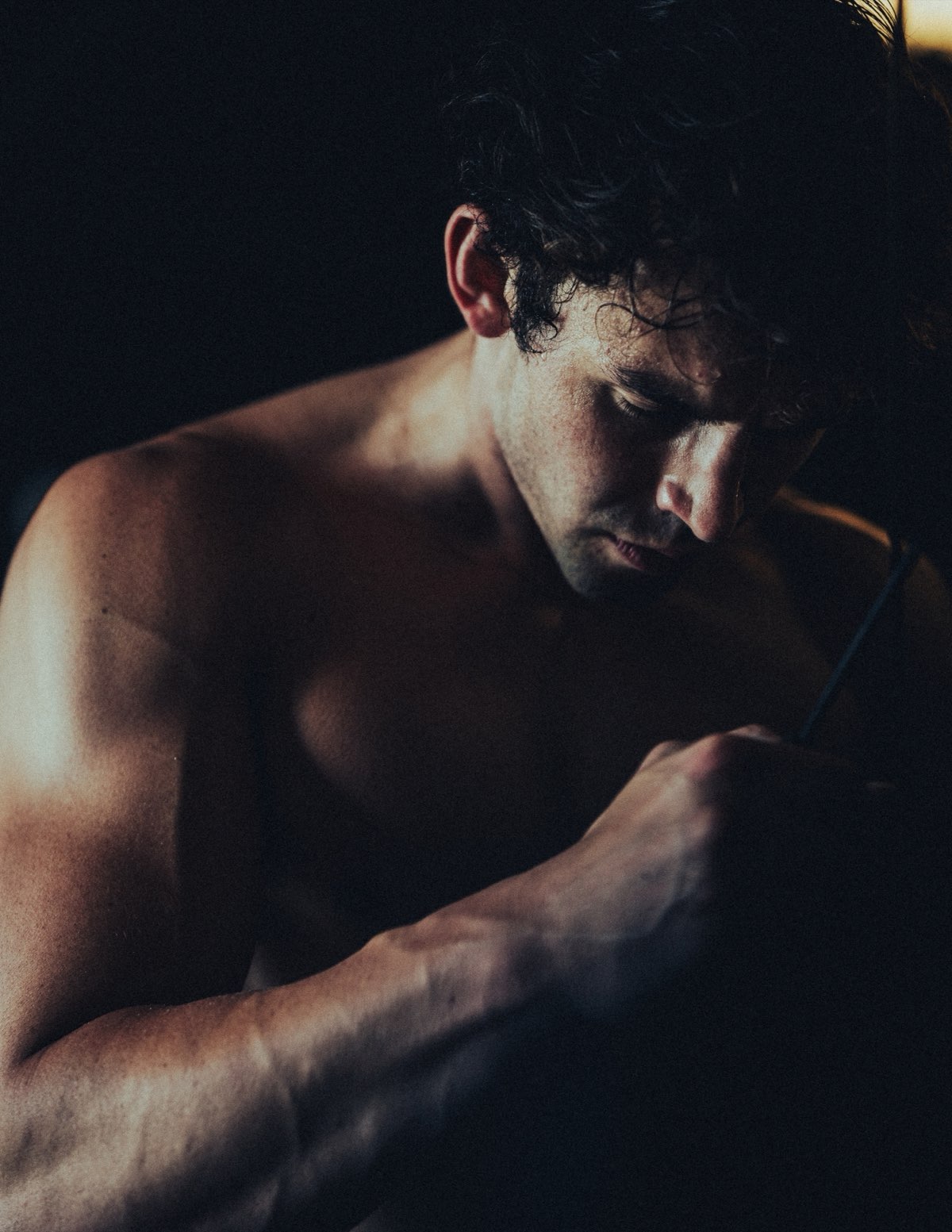 Anthony shirtless in low warm light, head down, hands working a grip